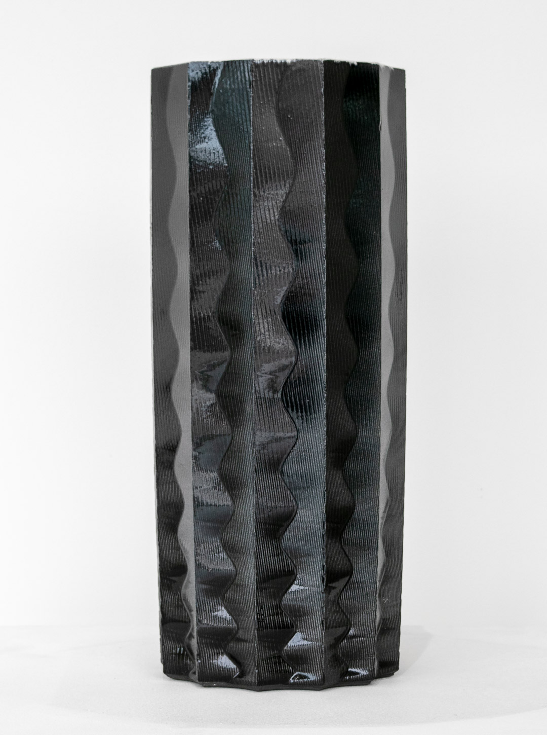 A cylindrical black porcelain vessel with vertical ribbed texture and geometric wedge-shaped cutouts creating negative space throughout. The sculptural form features alternating light and shadow created by the carved pattern, presented against a neutral white background.