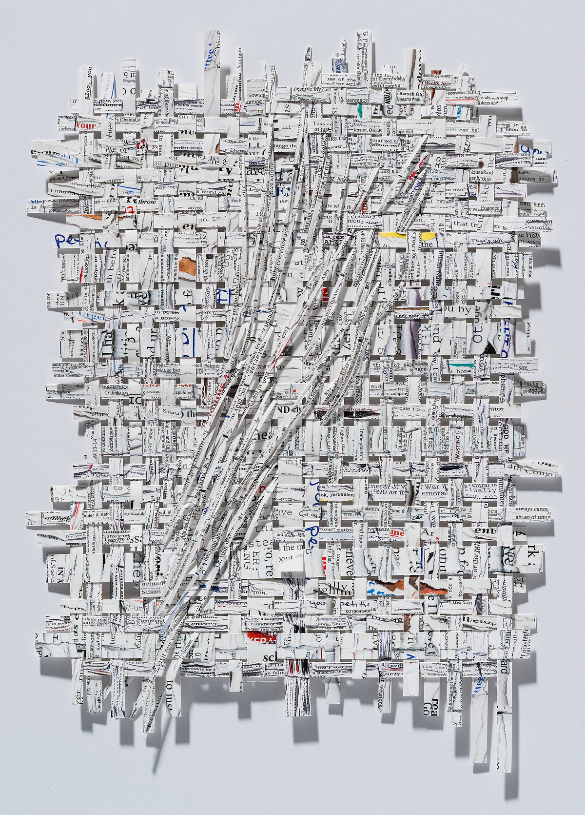 A three-dimensional collage of shredded and layered printed text and newspaper fragments arranged on a white wall, creating a dense, textured composition with diagonal striations and scattered edges. Occasional pops of color—including yellow, orange, red, and blue accents—punctuate the predominantly white and gray tones.