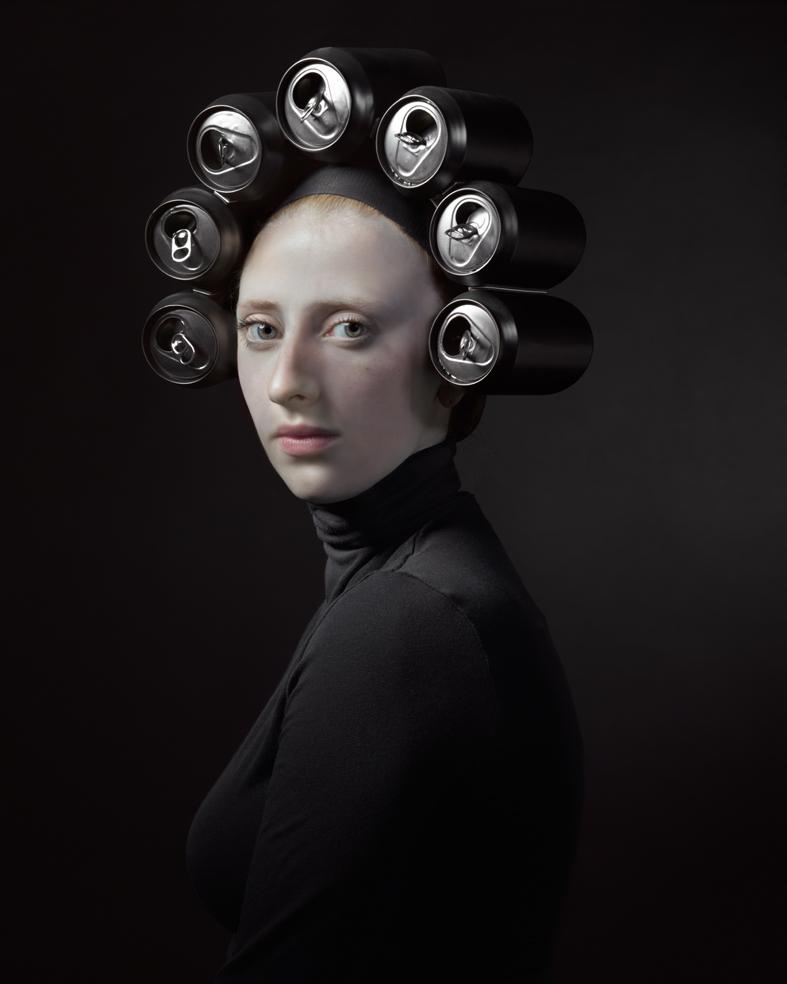 Portrait of a pale-faced figure in profile against a dark background, wearing a black turtleneck. Seven metallic beverage cans with pull-tab openings are arranged in a halo around their head, their reflective surfaces catching light. The stark, high-contrast composition combines classical portraiture with contemporary consumer materials.