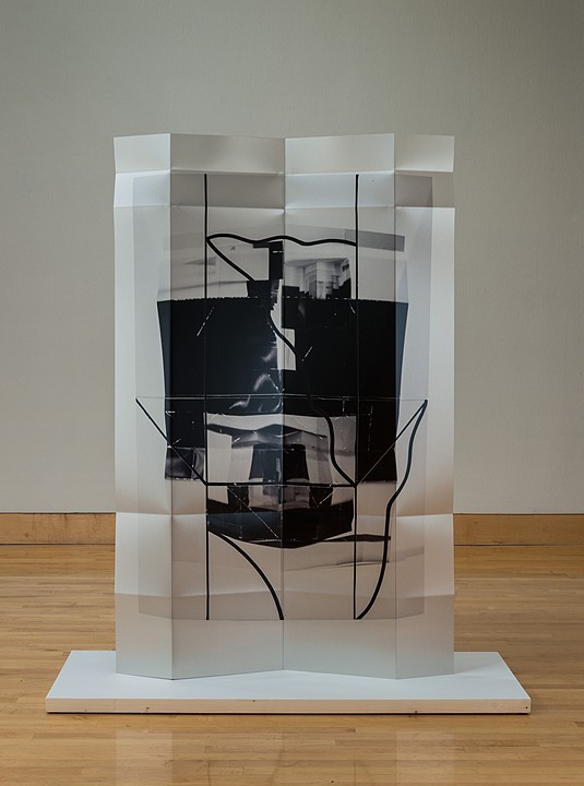 Translucent panels of frosted and clear material are arranged in a tall rectangular structure, layering to create depth. Within the center, bold black silhouettes of intertwining curved lines and organic shapes are visible through the stacked panels, creating a striking contrast against the light surfaces.