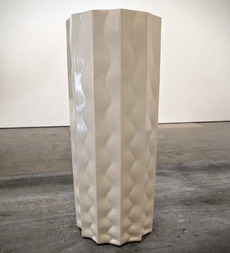 A tall, cylindrical white porcelain vase with a hexagonal cross-section stands on a concrete floor. The surface is decorated with repeating wavy vertical patterns in subtle relief, creating a quilted or undulating texture across all sides.