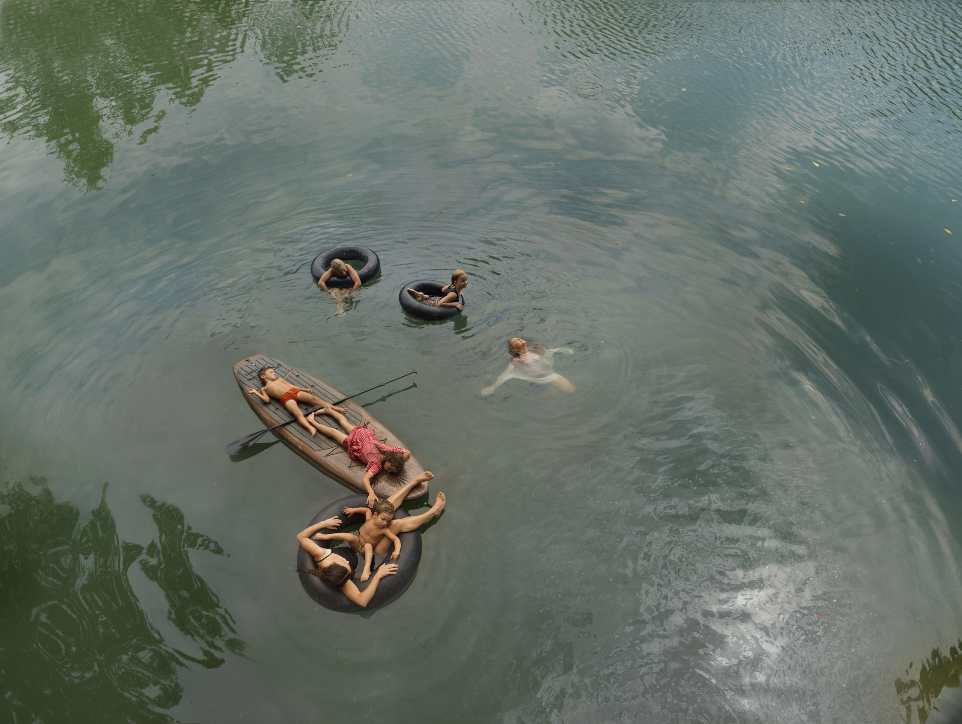Aerial view of people cooling off in murky water, some floating on inner tubes and a wooden raft, others swimming freely, surrounded by green vegetation at the water's edge.