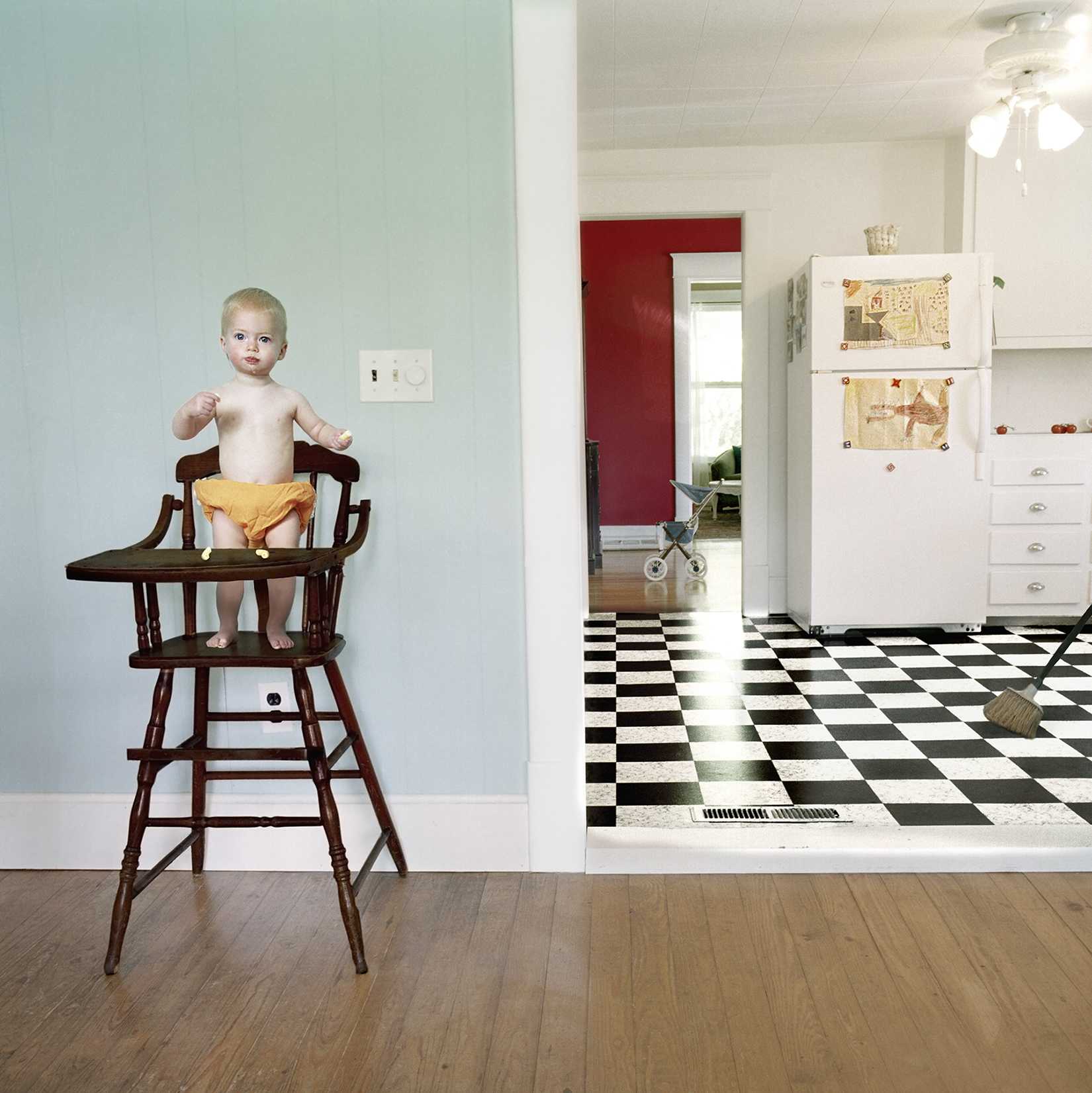 A toddler with blonde hair sits in a vintage wooden high chair wearing yellow pants, positioned in a light blue-walled room with wood flooring. Beyond an open doorway, a retro kitchen features black and white checkered flooring, white cabinetry, and a red door.