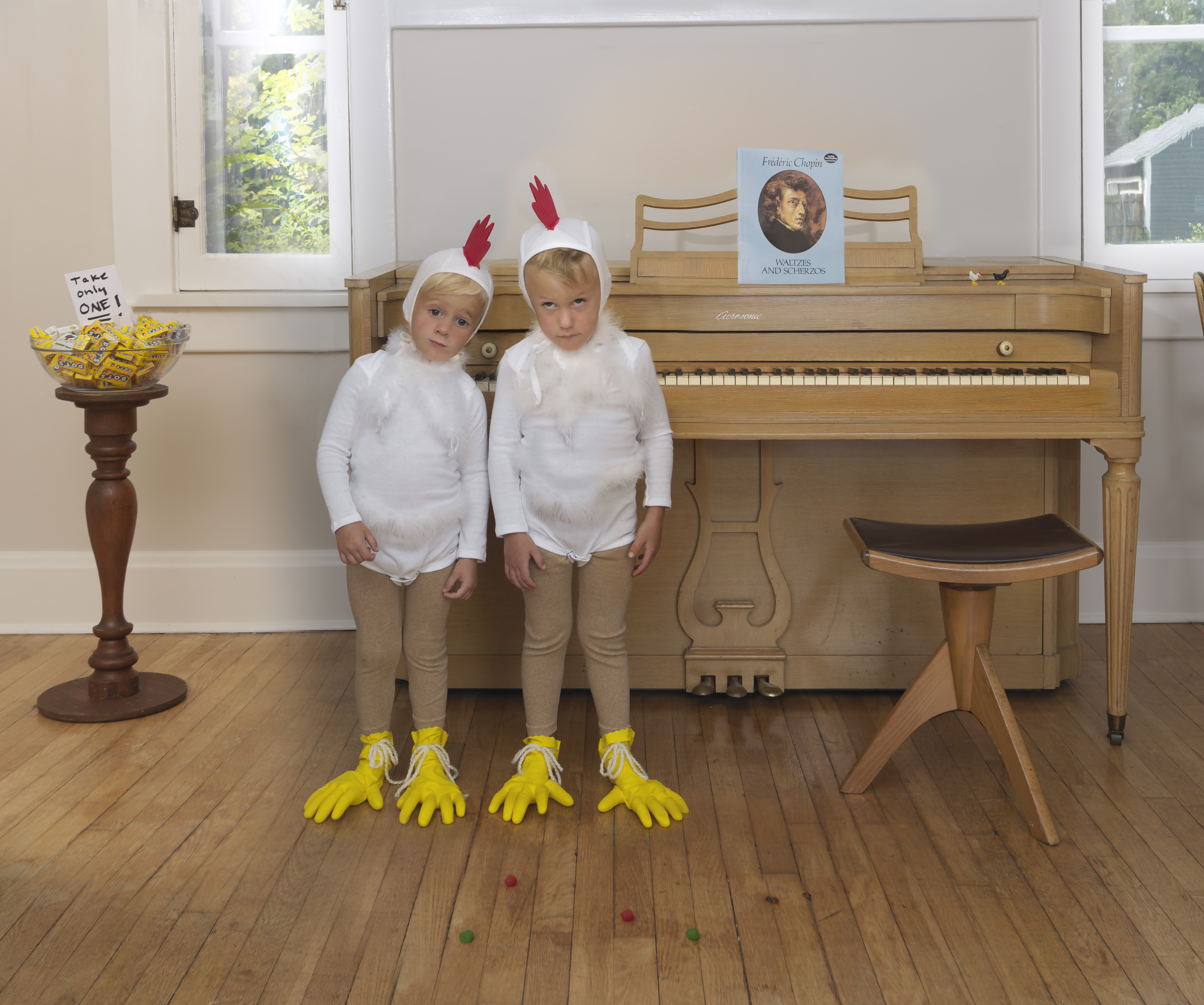 Two young children in white long-sleeved shirts and tan pants stand before a wooden upright piano, wearing white headbands with red feathers and oversized yellow gloves. Colorful balls are scattered across the wooden floor, while a classical music score rests on the piano's top. A pedestal table holding a bowl of candy and a sign reading "TAKE ONLY ONE" sits to the left, and large windows overlook greenery in the background.