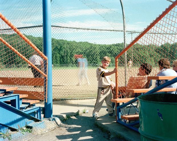A baseball dugout scene viewed through a chain-link fence, with players and spectators in a sunny outdoor stadium. Vibrant primary colors—bright blue structural elements, orange and red netting, and green equipment—contrast against the natural tan infield and tree-lined background.