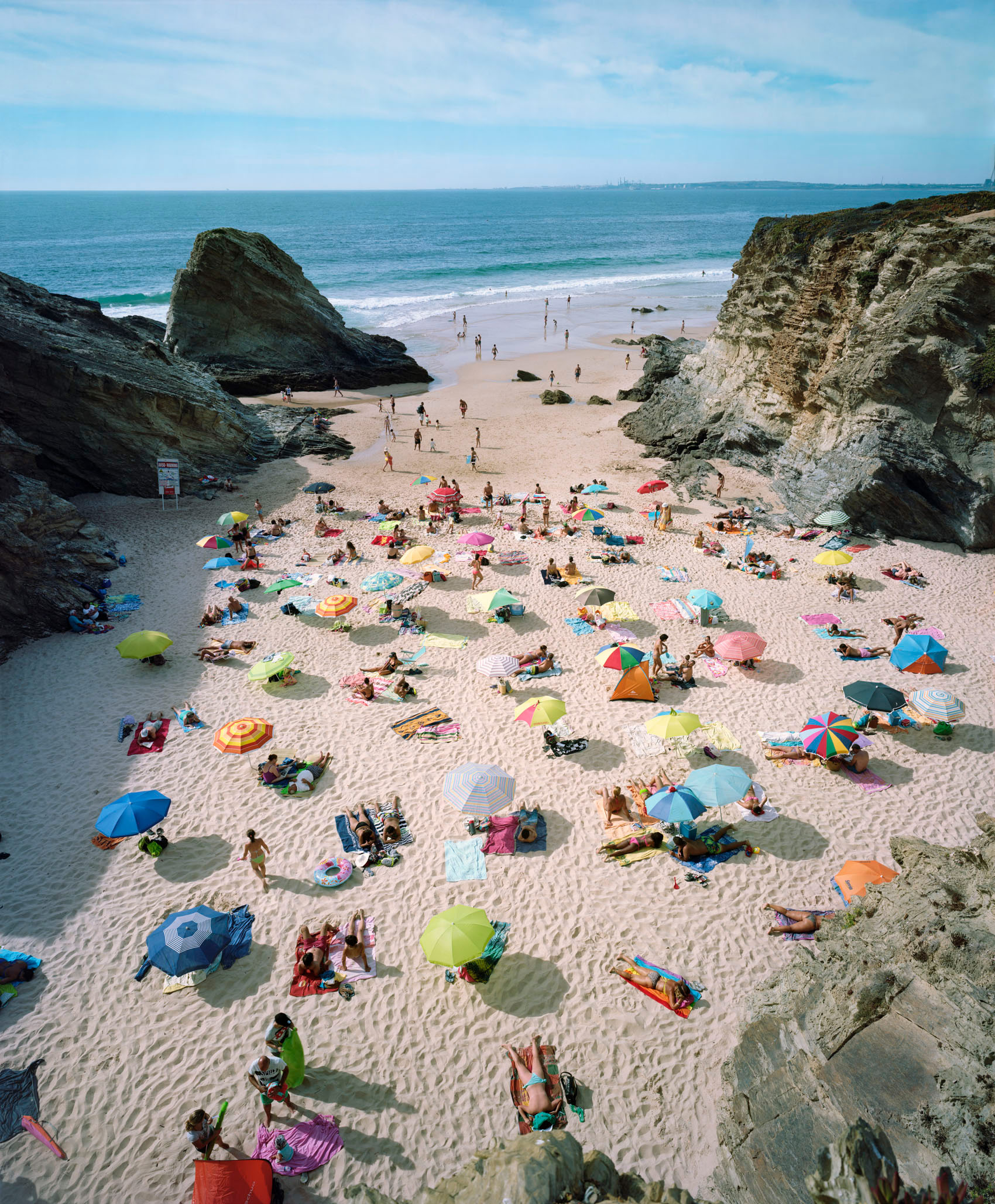 Aerial view of a crowded beach nestled between dramatic rocky cliffs, with numerous colorful umbrellas and sunbathers scattered across the sand. The composition captures the vibrant contrast between the natural landscape and the bright blues, pinks, yellows, and greens of beach equipment dotting the golden shore.