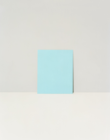 A light blue rectangular form stands vertically against a neutral beige wall and floor, casting a subtle shadow. The minimalist composition emphasizes the geometric shape and its interaction with the surrounding space.