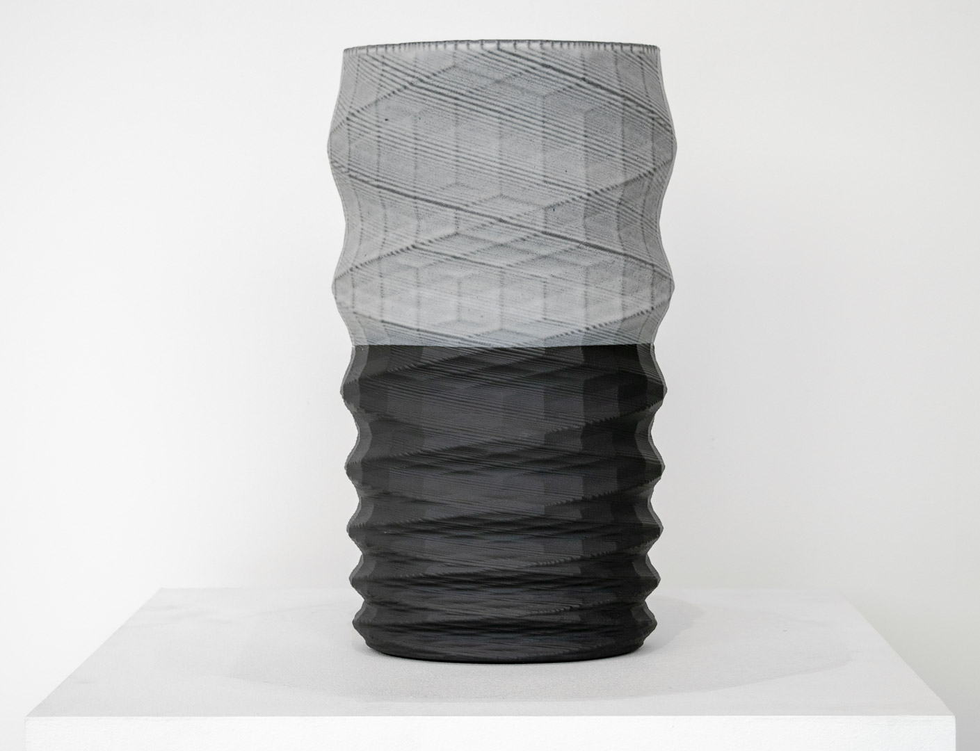 A tall cylindrical vessel with a twisted, ribbed surface sits on a white platform. The upper half features a translucent gray mesh-like texture, while the lower half is rendered in solid black with horizontal ridging, creating a striking color and material contrast.