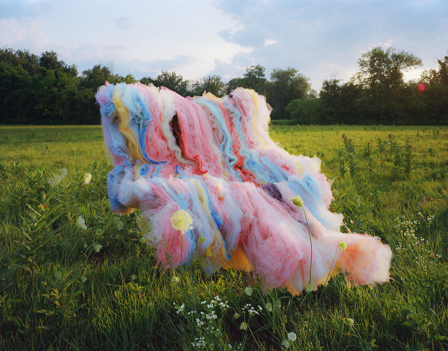 Voluminous layers of colorful tulle fabric in shades of pink, blue, yellow, and white billow across a green meadow dotted with wildflowers, creating an ethereal sculptural form against a cloudy sky and tree line.