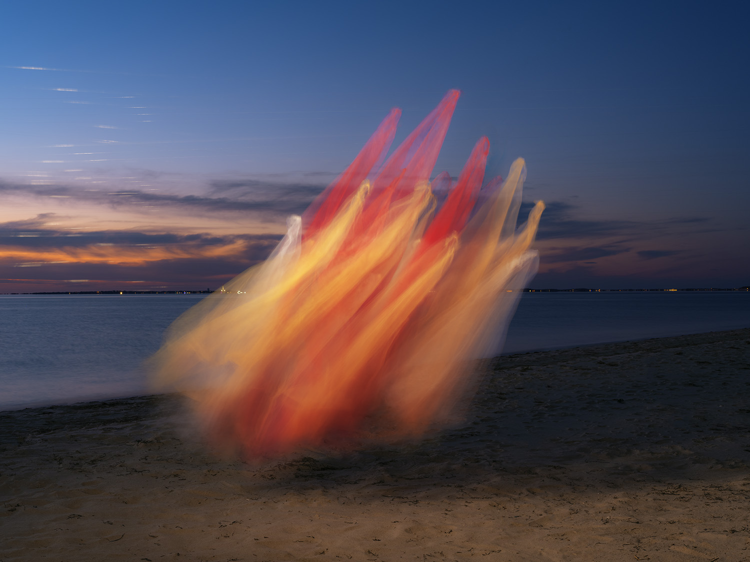 A dynamic sculptural form composed of billowing red, orange, and cream-colored fabric captured mid-motion against a twilight sky. The translucent tulle sweeps upward from a beach in dramatic angular folds, with water and distant shoreline lights visible behind it.