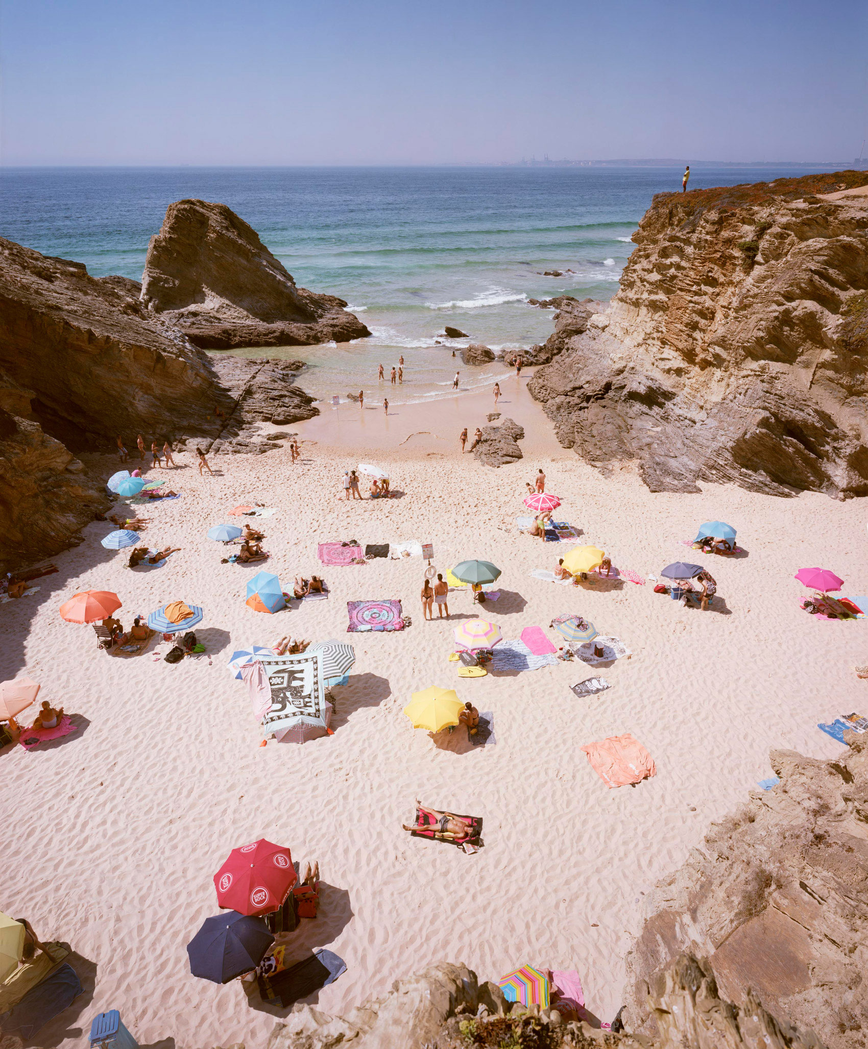 Aerial view of a crowded beach cove enclosed by dramatic white limestone cliffs, with scattered beachgoers beneath colorful umbrellas in red, pink, yellow, and blue scattered across pale sand, calm turquoise waters visible beyond.