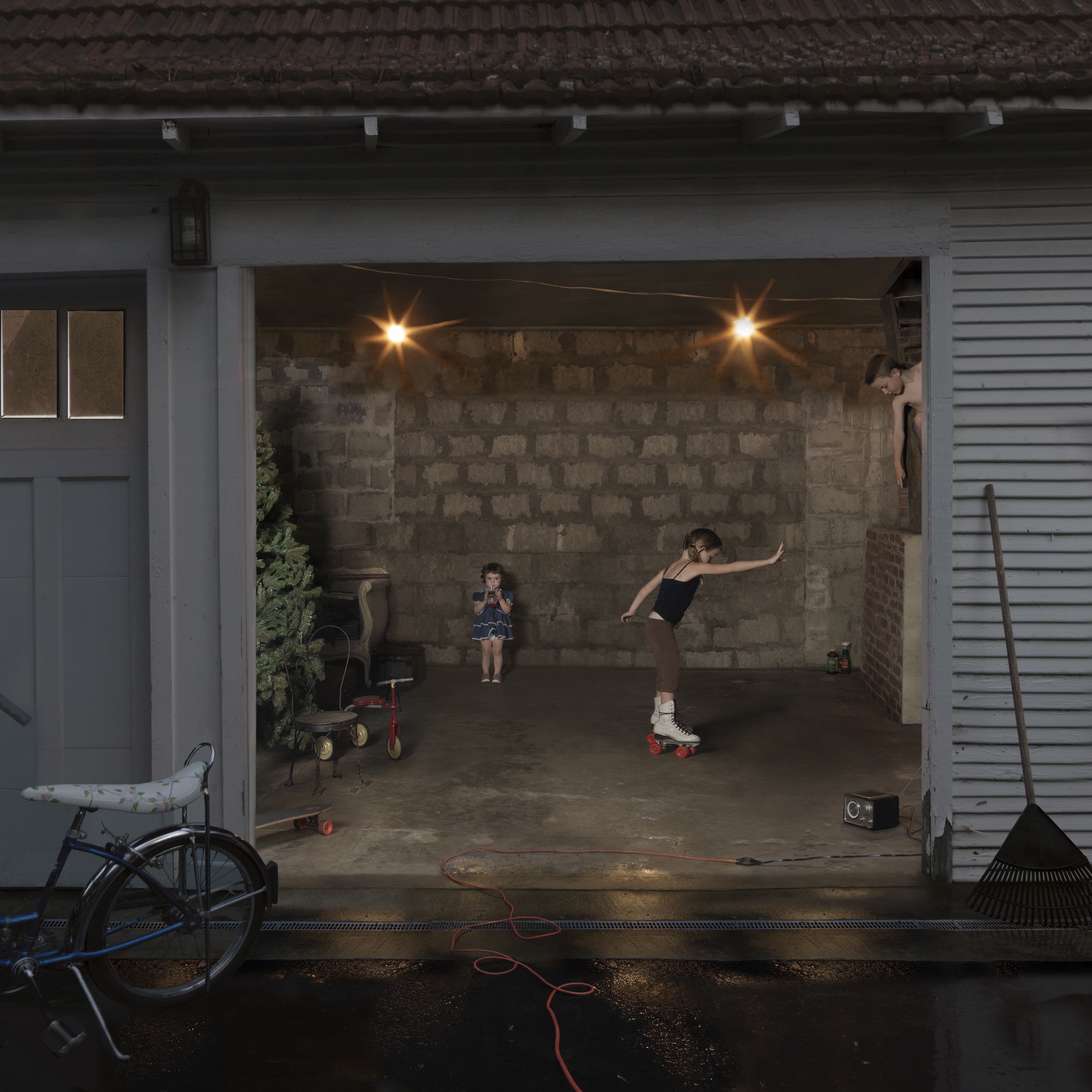 A garage interior photographed at dusk shows two children engaged in play: one wearing roller skates performs a dynamic pose on the concrete floor while another stands nearby in a blue dress. The space is illuminated by two overhead lights, revealing exposed cinder block walls, various toys and household items scattered about including a tricycle and bicycle, and tools leaning against the sides. The photograph captures an intimate domestic moment framed by the open garage door.