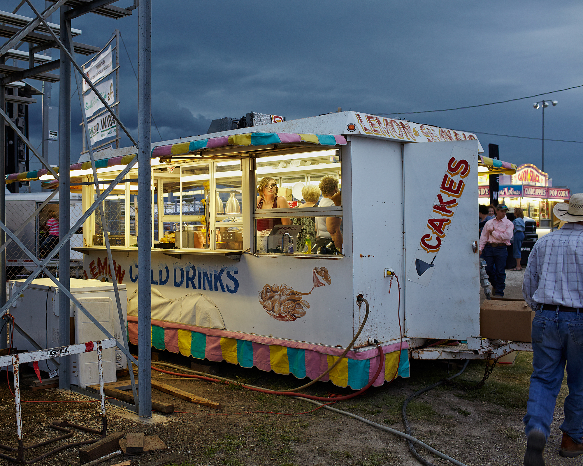 A vintage concession stand with white exterior walls and colorful striped awning in pink, yellow, and turquoise trim sits at what appears to be a county fair or carnival. Text on the stand reads "CAKES" and "COLD DRINKS" with decorative pretzel imagery. Several customers stand outside the service window where staff members are visible inside, with fairground infrastructure and other vendor booths visible in the background under a dramatic cloudy sky.
