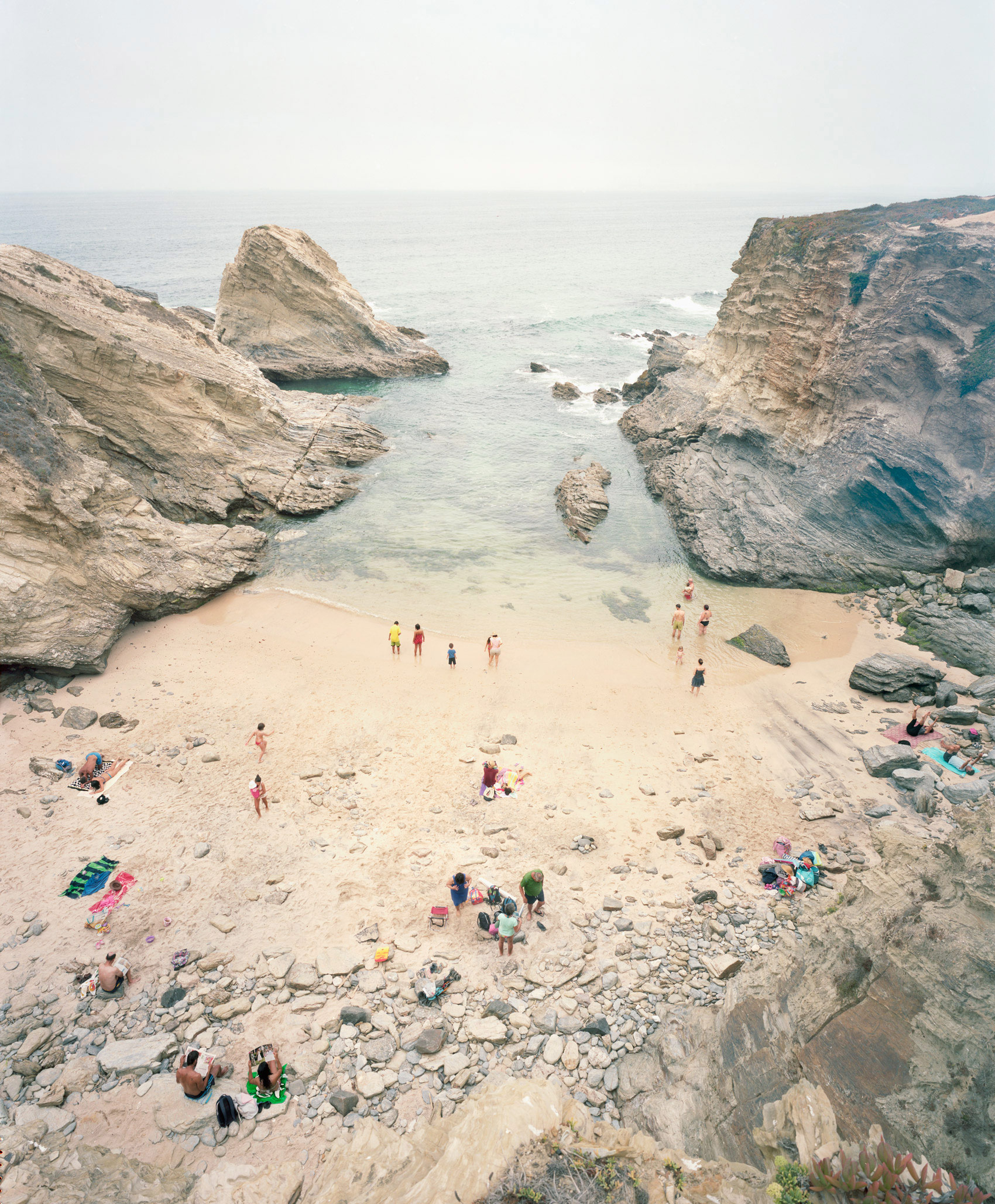 Aerial view of a secluded sandy cove framed by towering stratified limestone cliffs, with scattered beachgoers in colorful clothing and swimwear dispersed across the beach and shallow turquoise waters below.