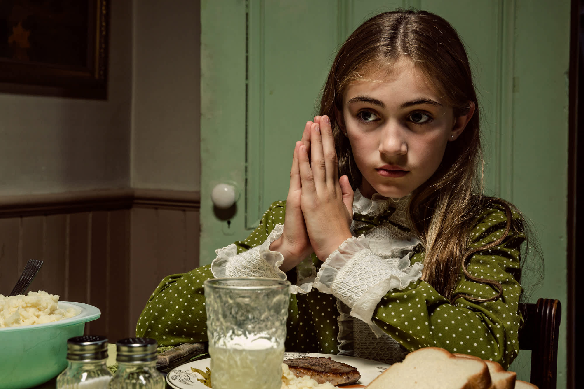 A young girl in a olive-green polka dot dress with white lace cuffs sits at a dining table with her hands pressed together near her face in a gesture of prayer or contemplation. Before her are a glass of water, a plate with bread, and a bowl of food, set against pale green walls in an interior domestic scene rendered in rich, saturated color.