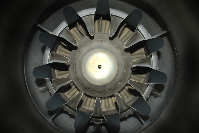 Overhead view of a circular mechanical structure with black blade-like forms radiating outward from a pale central hub, photographed against a dark background. The composition emphasizes radial symmetry and industrial materiality, with metallic and matte surfaces creating subtle tonal contrasts.