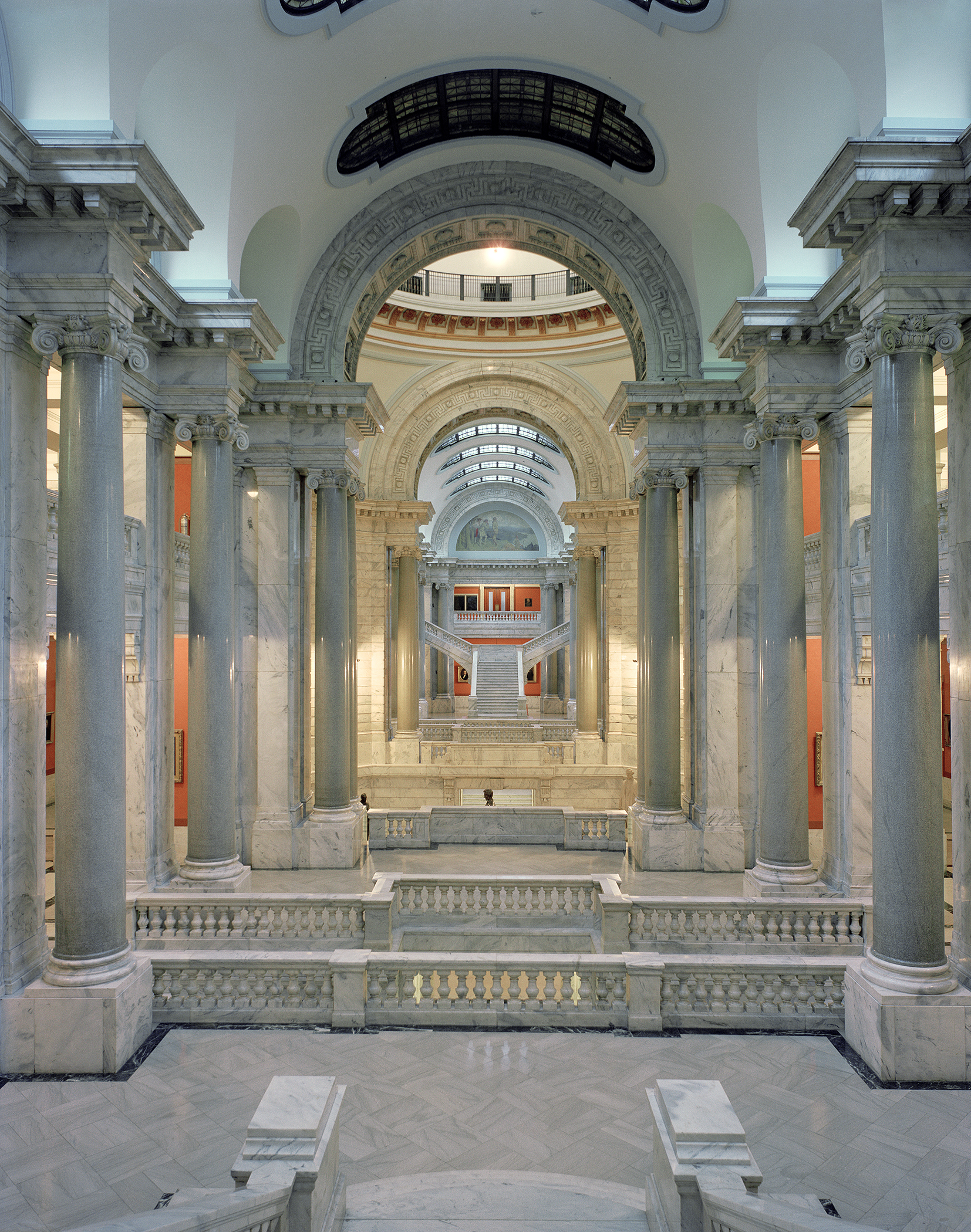 A symmetrical corridor with towering fluted columns in pale marble flanks a central passage, leading to a grand arched rotunda with ornamental detailing. Warm lighting illuminates the neoclassical architecture, while burnt orange accent panels and ornate balustrades recede into the distance.