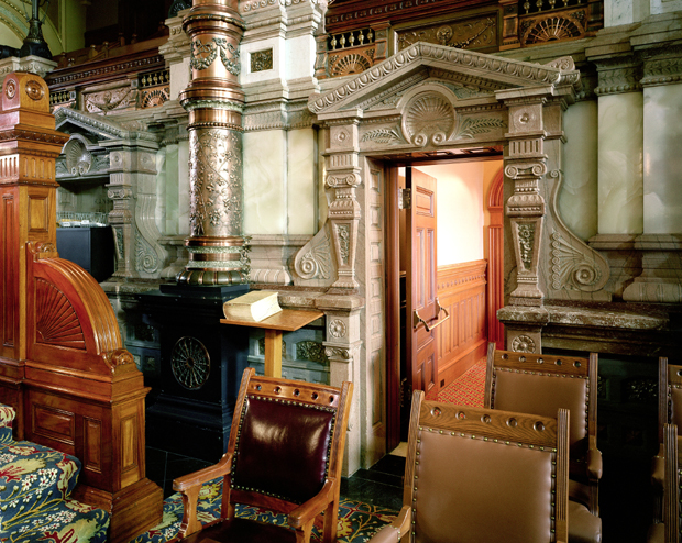 Interior view of an ornately decorated Victorian room featuring elaborate carved stone columns, decorative moldings, and cornices in cream and gilt tones. The space contains period furnishings including a burgundy leather chair, wooden furniture, a black cast-iron stove, and a red-orange wooden door, with patterned flooring visible in the foreground.