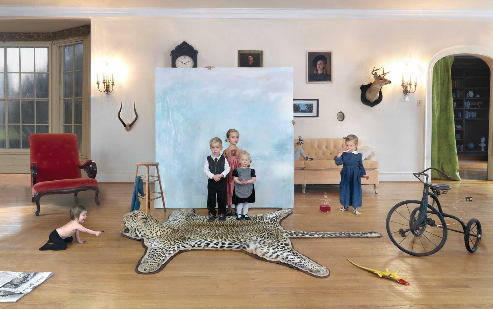 A staged interior scene featuring five children of varying ages positioned throughout an elegant room with hardwood floors. Children are arranged among surreal domestic elements including a large spotted leopard skin rug, a red velvet chair, a vintage bicycle, a tan sofa, and a large canvas painting depicting three of the children against a pale blue background. The room is decorated with taxidermied animal heads, wall sconces, framed portraits, and a clock, creating an absurdist juxtaposition of refined décor and playful childhood chaos.