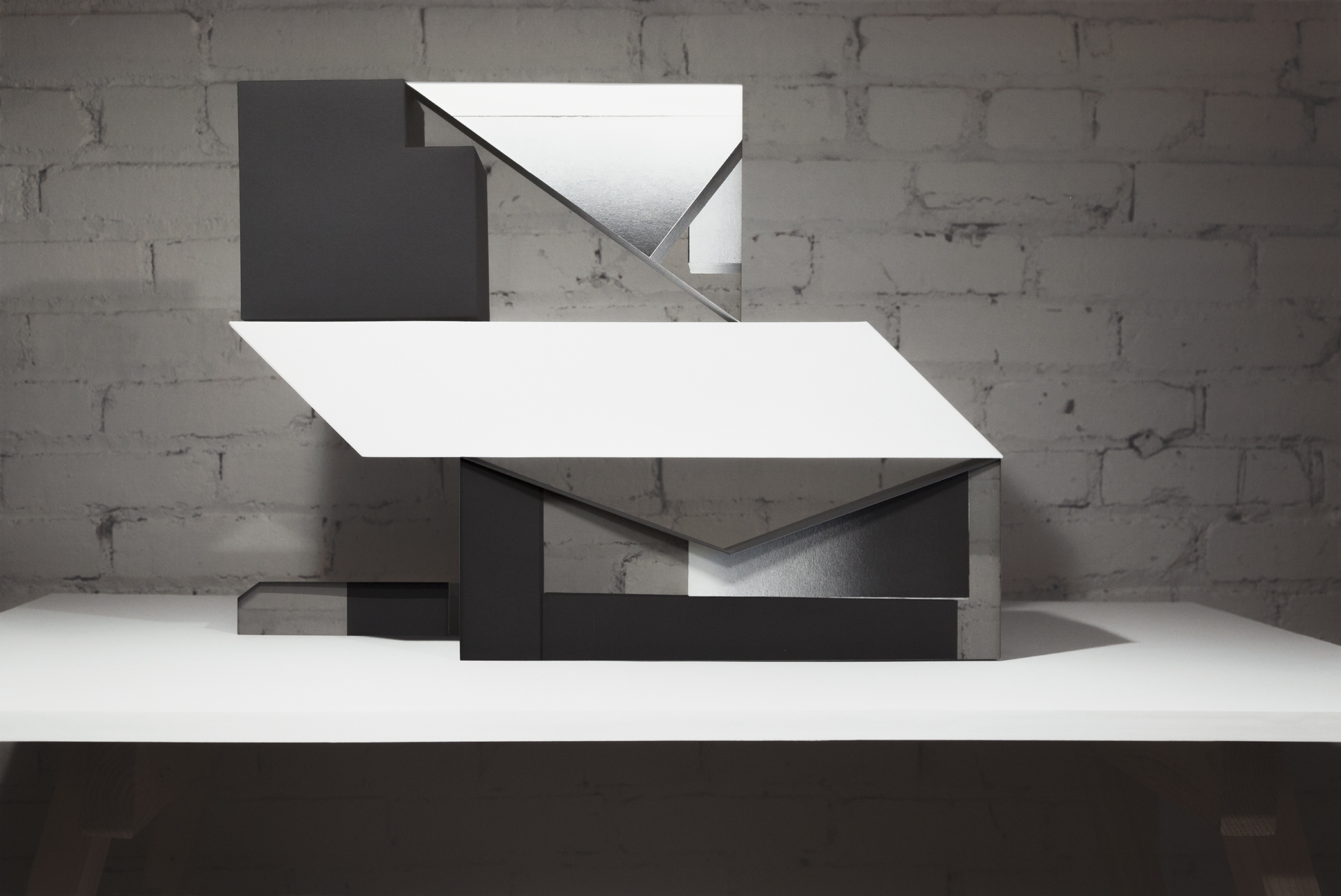 An abstract three-dimensional sculptural form constructed from layered white and black materials, displayed on a white platform against a weathered brick wall. The composition features angular, intersecting planes with folded and cut surfaces that create dynamic shadows and spatial depth.