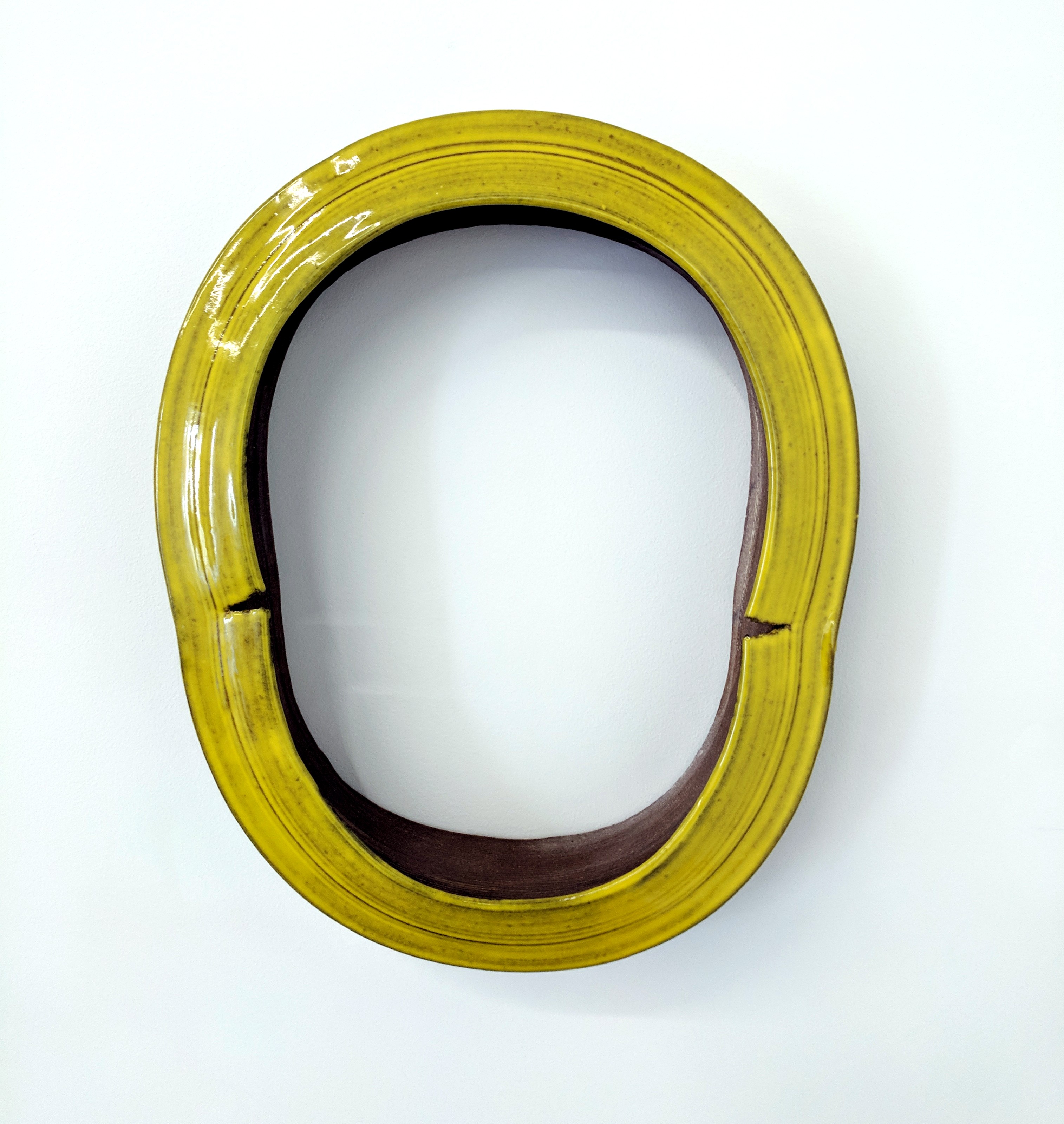 A vibrant yellow-green glazed ceramic ring form with an oval opening, set against a white background. The glossy surface contrasts with the darker interior, and subtle linear textures run across the frame's width.