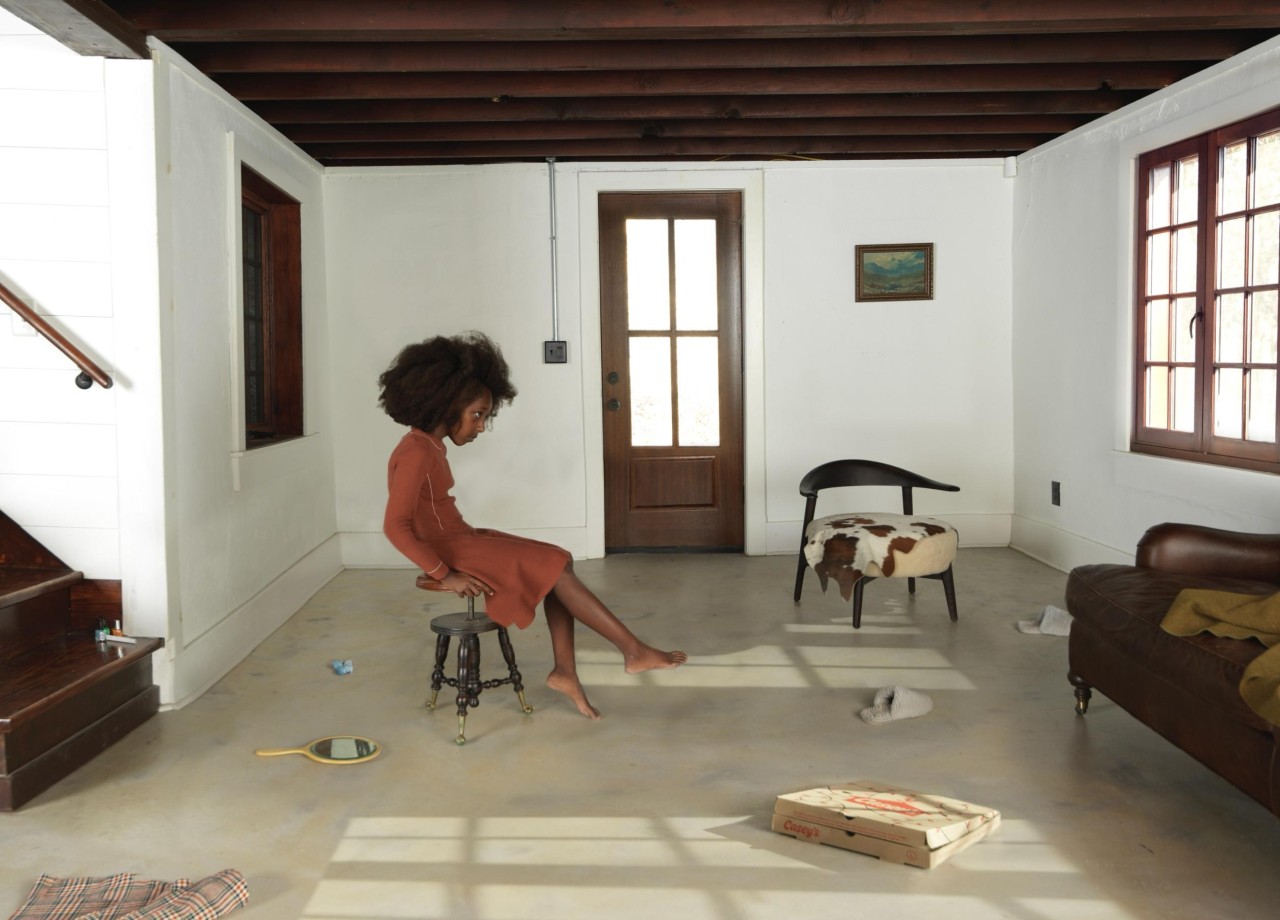 A woman in a rust-colored dress sits on an industrial stool in a sparse, sunlit interior space with white walls and dark wooden beams. The minimalist room contains scattered objects including books, a patterned rug, and modernist furniture, with natural light casting geometric shadows across the polished concrete floor.