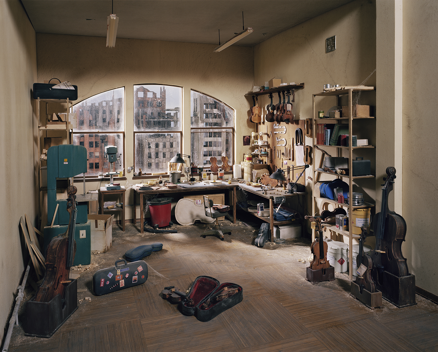 A meticulously arranged workshop interior featuring multiple workbenches, wooden shelving units, and numerous string instruments in various states of repair. Tools hang organized on pegboards, while violin cases and instrument parts are scattered across the floor and tables. Large arched windows overlook an urban cityscape of brick buildings, and fluorescent and pendant lighting illuminates the neutral-toned walls and wooden flooring.