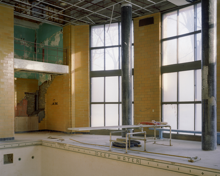 Abandoned indoor swimming pool facility with yellowed tile walls, a deep water measurement marker visible along the pool edge, industrial metal support columns, large gridded windows flooding the space with natural light, and scattered construction materials on the empty pool floor.