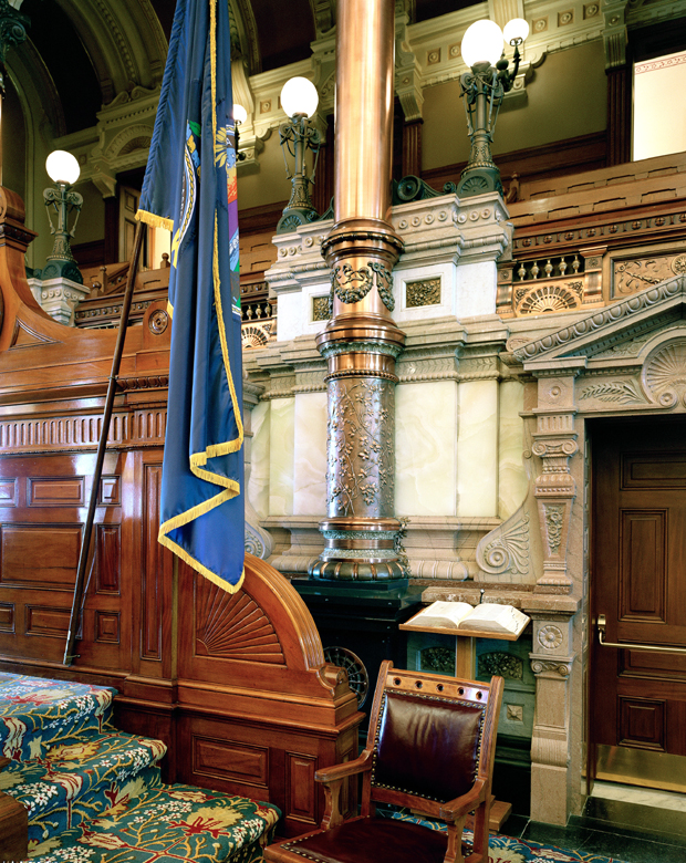 Interior view of an ornately decorated government building chamber featuring classical architectural elements including carved columns, decorative molding, and coffered ceilings. A blue flag with gold trim hangs prominently on the left, while wood paneling, brass light fixtures, and period furnishings in jewel tones occupy the foreground.
