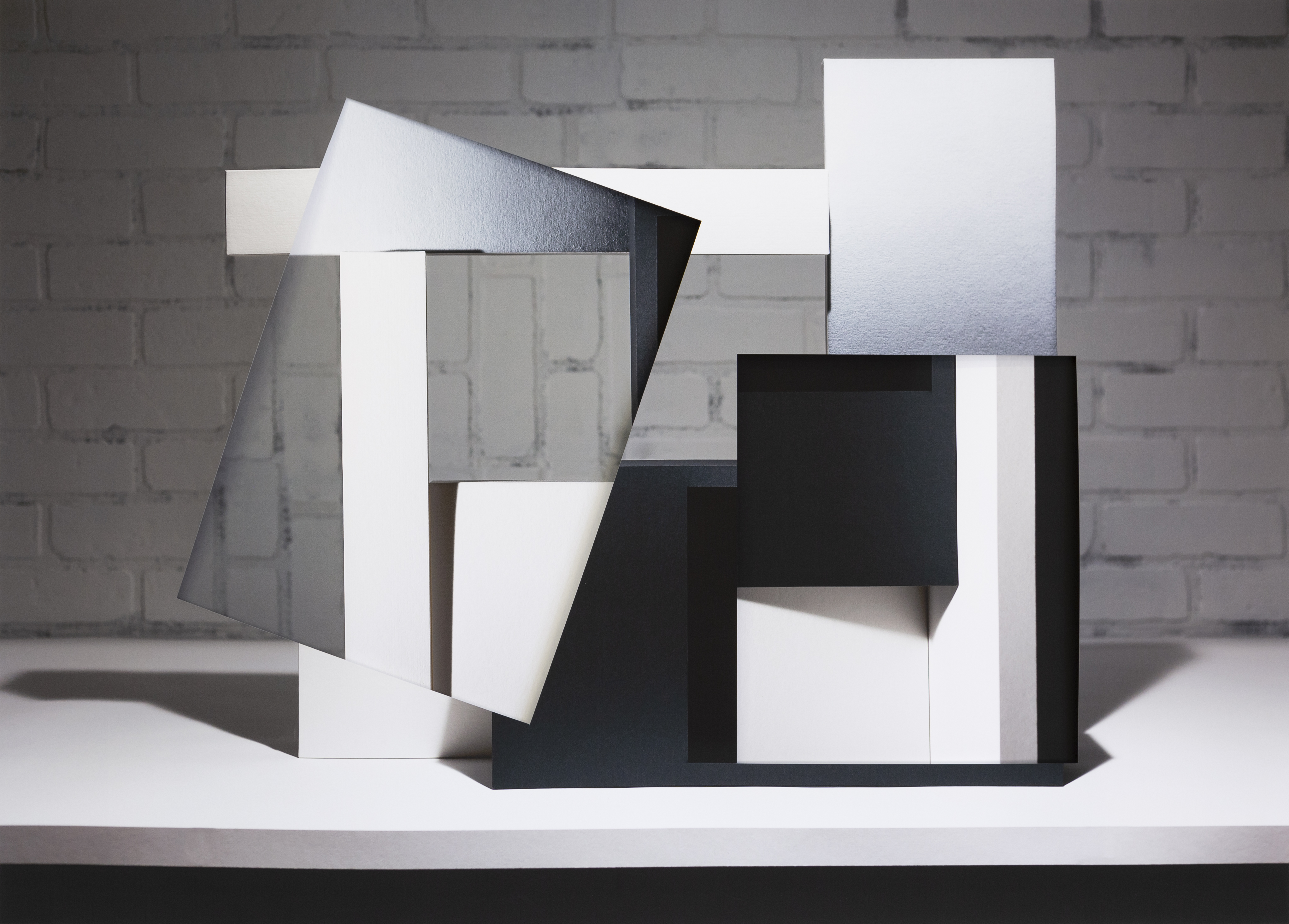 Abstract collage of white and black geometric forms layered and overlapped against a white brick wall backdrop. The composition features cut and folded paper planes creating sharp angles and shadows, with varied surface textures ranging from glossy to matte finishes, arranged in a three-dimensional sculptural arrangement on a white platform.