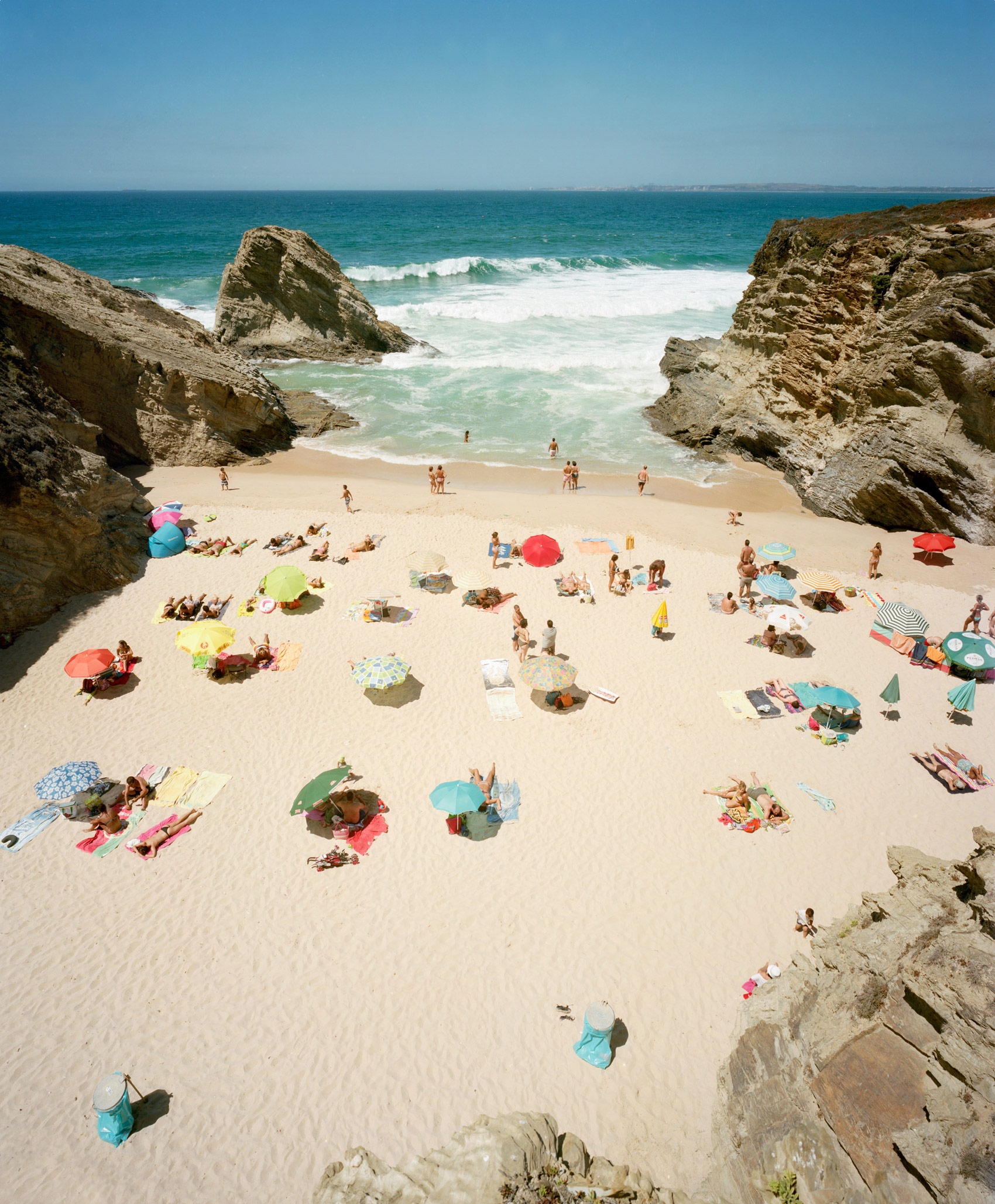 Aerial view of a secluded beach cove framed by dramatic limestone cliffs, with dozens of beachgoers scattered across pale sand beneath colorful umbrellas and beach shelters in shades of red, pink, yellow, blue, and green, turquoise waves visible beyond.
