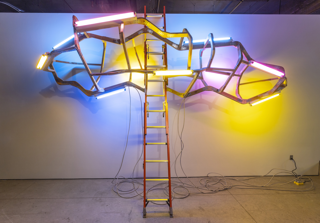 Neon light sculpture mounted on an extension ladder featuring geometric wooden frames in yellow, blue, and magenta, with illuminated tubes creating a symmetrical, wing-like composition against a gallery wall.