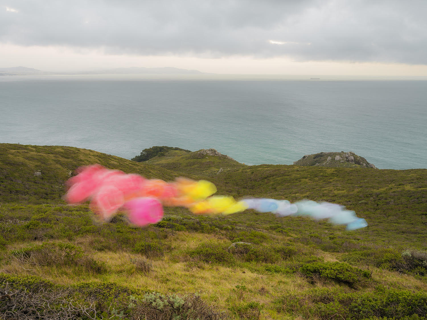 Blurred rainbow-colored tulle fabric streaks across a green coastal hillside overlooking turquoise ocean waters under an overcast sky, creating an ethereal contrast between the delicate, translucent material and the rugged landscape.