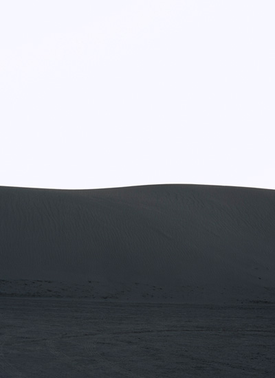 Minimalist landscape with a pale gray sky above a dark charcoal horizon line, beneath which lies a textured darker plane suggesting water or terrain, creating a stark division of tones and planes across the composition.