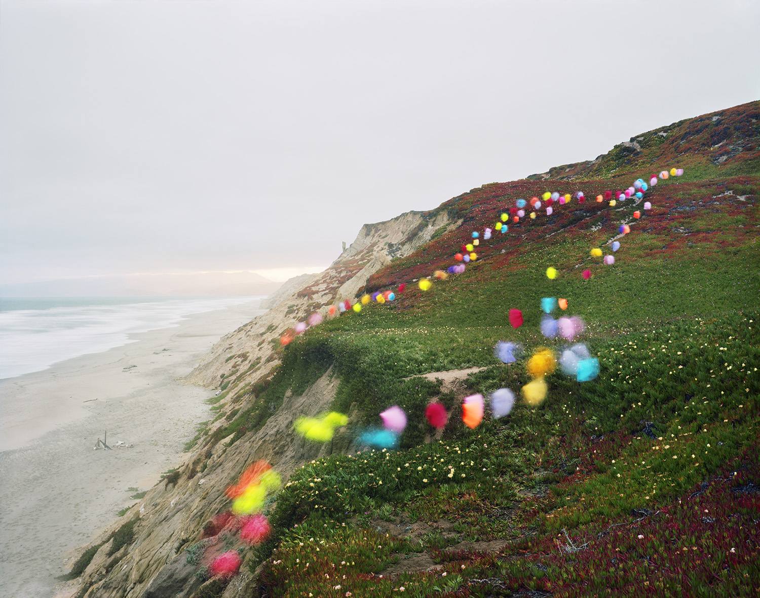 Colorful inflated spheres in shades of pink, yellow, blue, cyan, and red are arranged in curved lines across a coastal hillside covered in green vegetation and rust-colored plants. The spheres create a playful path descending toward a dramatic eroded cliff face and sandy beach below, with calm ocean visible in the distance.