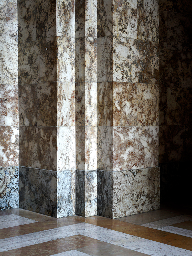A corner view of a geometric interior featuring alternating vertical stripes of weathered white and dark surfaces, with distressed textures and rust-colored staining. The composition shows layered planes meeting at a right angle, with a polished floor in warm and cool tones reflecting the striped wall treatment.