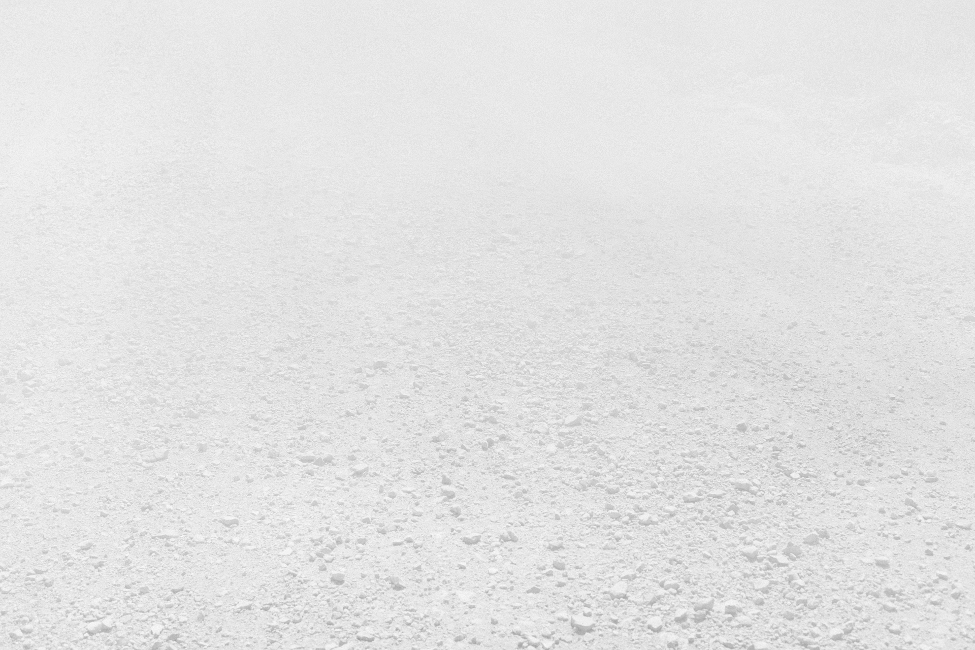 A vast, minimalist landscape rendered in pale gray and white tones, depicting an expansive flat terrain with subtle variations in texture and density across the surface. The composition recedes toward a hazy horizon, creating an immersive sense of emptiness and atmospheric perspective.