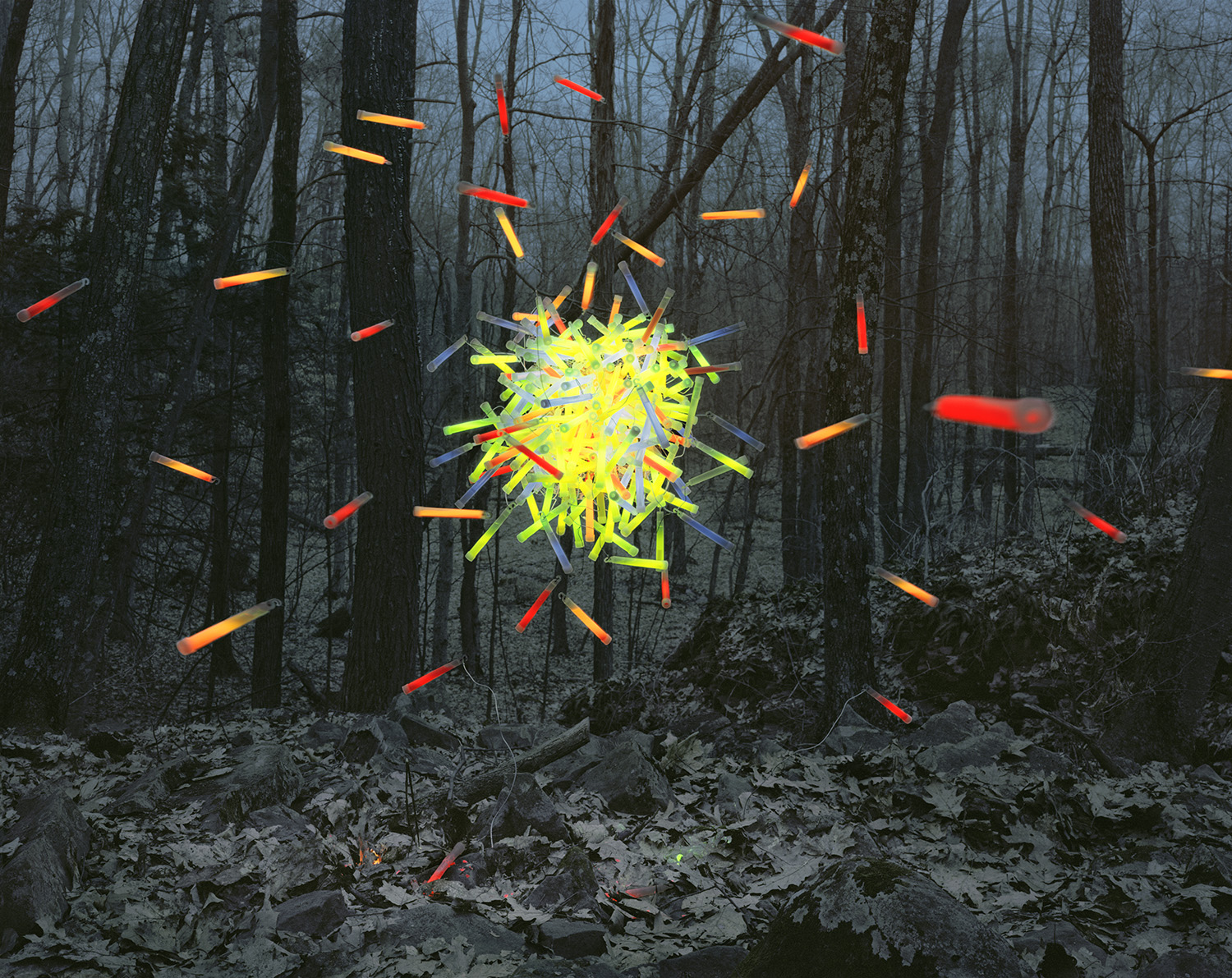 A dense forest of bare trees frames a radiant burst of neon yellow and multicolored glowing sticks exploding outward from the center, with additional colored sticks in red, orange, and yellow scattered throughout the darker woodland scene.