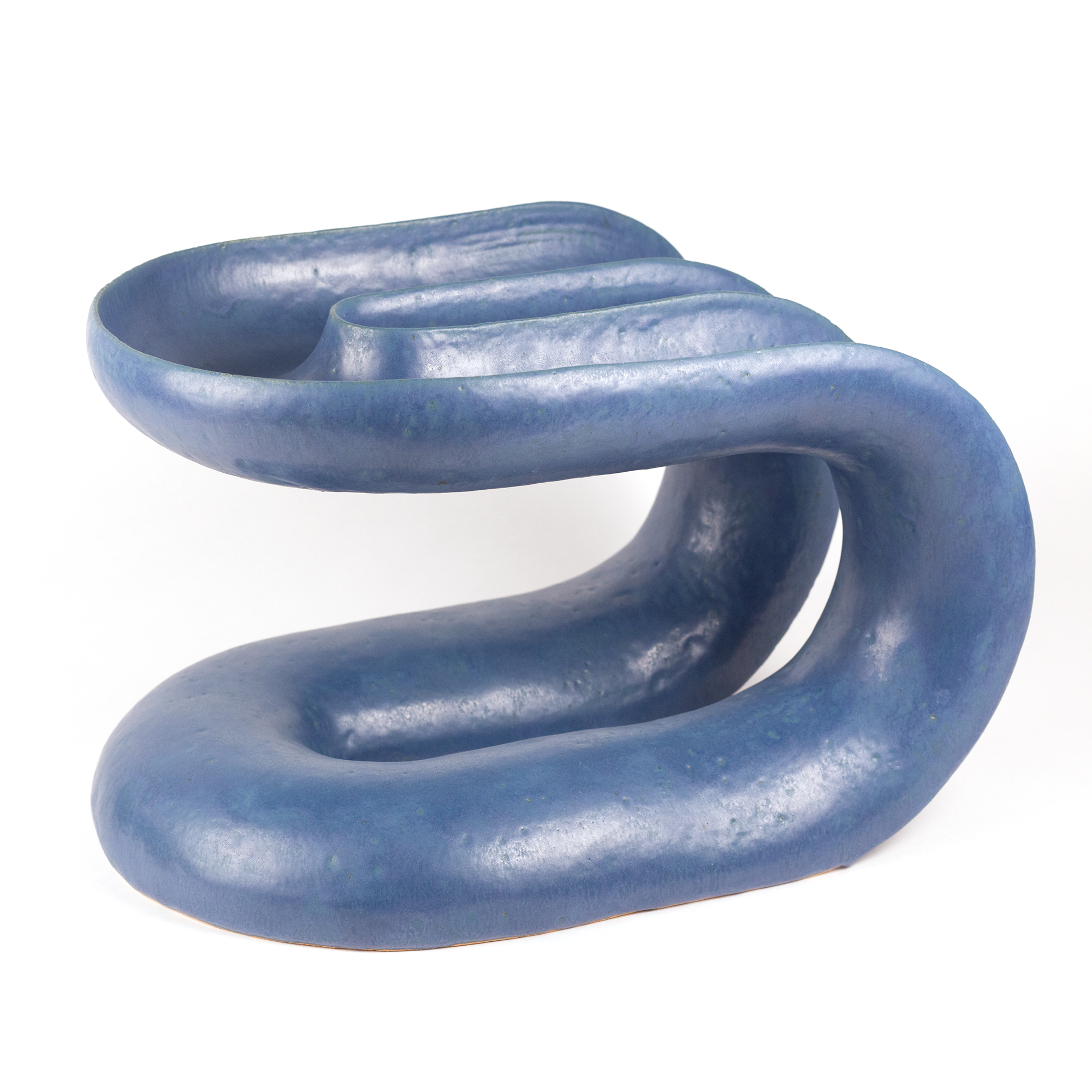 A sinuous blue ceramic form curves and loops organically, with smooth glazed surfaces that shift between darker and lighter tones. The sculptural object twists in on itself, creating multiple hollow chambers and openings that invite the viewer to observe it from various angles.