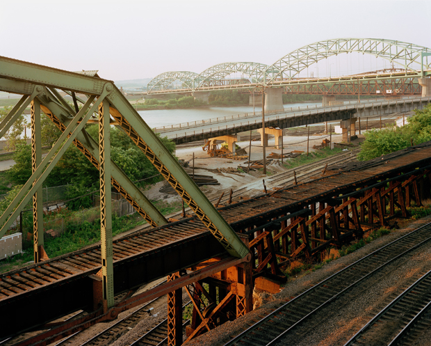 Industrial railroad bridge infrastructure in the foreground with rusted steel trusses and support beams, extending toward a modern arched bridge crossing a wide river in the distance. The composition layers deteriorating metalwork, active rail yards with equipment and structures, and waterway landscape under overcast sky, rendered in muted greens, browns, and grays.