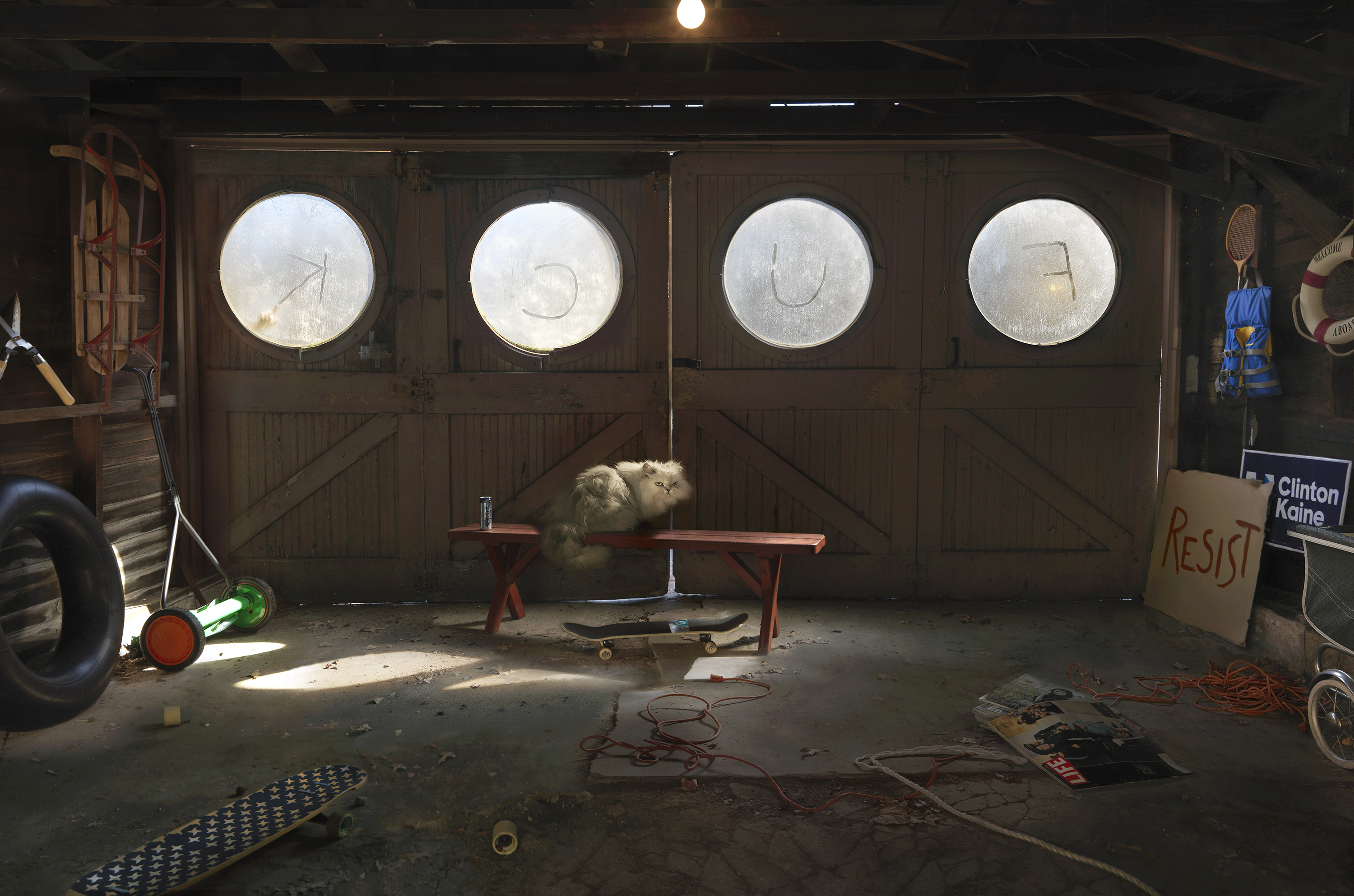 A cluttered garage interior featuring four large circular windows with frosted glass panes set into brown wooden doors. A fluffy white cat sits on a red bench beneath the windows. The concrete floor is scattered with miscellaneous objects including bicycle parts, coiled red cable, a skateboard, sports equipment, and political signage. Sunlight streams through the windows, creating bright geometric patterns across the space.