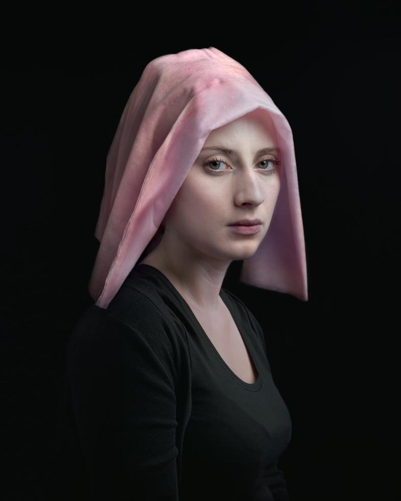 Portrait of a young woman in profile against a black background, wearing a black long-sleeved top and a draped pink fabric covering her head. Her pale complexion and direct gaze create a contemplative mood, with soft lighting emphasizing the delicate folds of the translucent cloth.