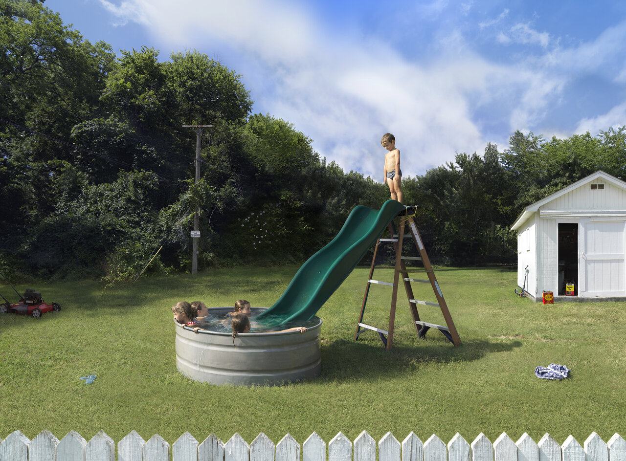 Children play on a slide attached to a large metal basin filled with water in a residential backyard, while another child stands atop the slide. A white shed, utility pole, mowed lawn, and dense green trees surround the scene under a partly cloudy sky, with a white picket fence visible in the foreground.