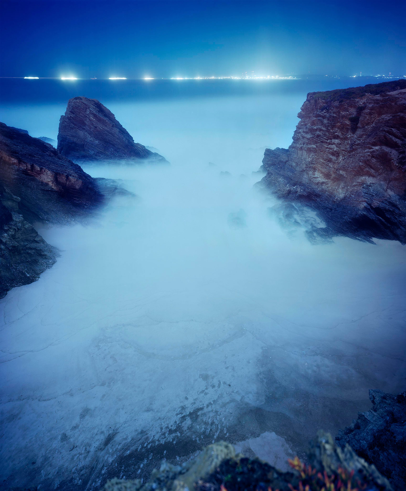 Twilight seascape with two rocky outcroppings framing a misty cove, their dark silhouettes contrasting against soft blue-white water and sky. A distant line of lights glows across the horizon in cool tones, creating a serene, long-exposure nocturnal composition.