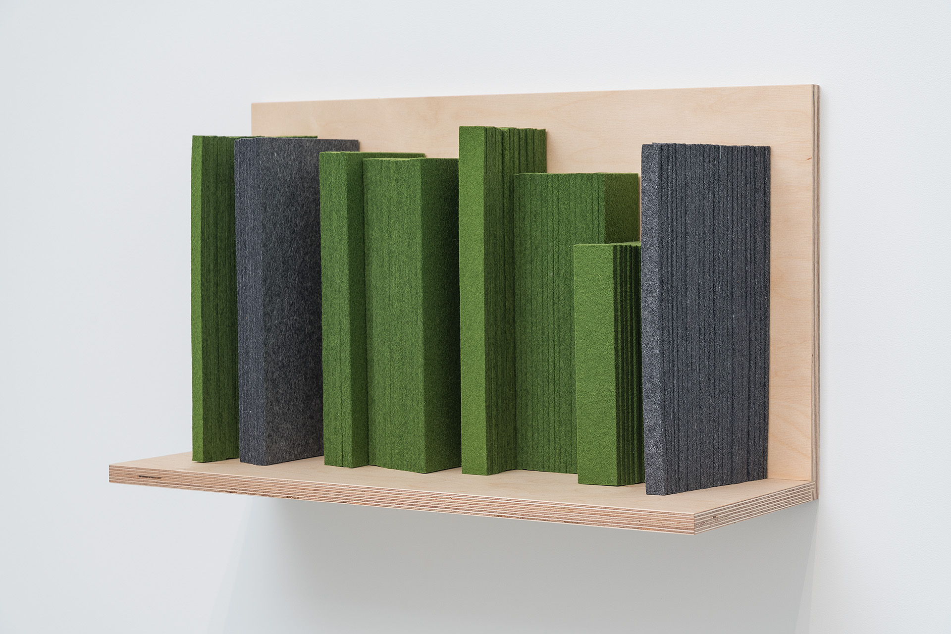 A wooden shelf mounted on a white wall displays nine vertical rectangular forms wrapped in textured felt in shades of green and grey, arranged in varying heights to create an abstract cityscape-like composition.
