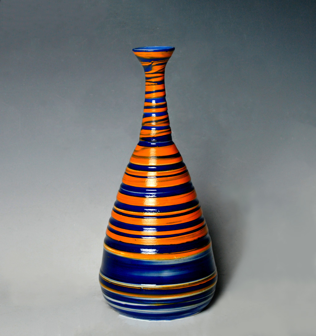 A tall conical ceramic vessel with a narrow neck flares dramatically toward the base, decorated with alternating horizontal stripes of vibrant orange and deep blue throughout. The glossy glazed surface reflects light, emphasizing the rhythmic band pattern that wraps continuously around the form.