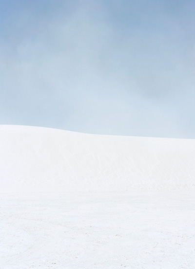 A minimalist landscape divided horizontally into two zones: a soft blue-gray sky occupying the upper portion, and a vast expanse of white snow or sand below, with subtle variations in tone creating gentle undulations across the lower plane.