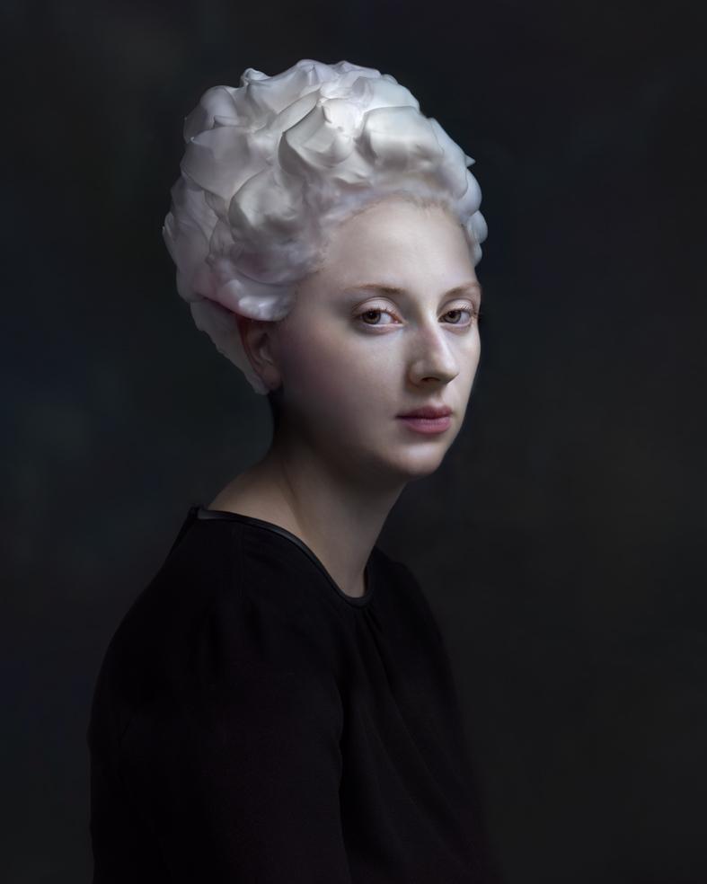 Portrait of a person with pale skin and white sculptural hair styled in voluminous waves, wearing a black garment against a dark background. The lighting is soft and dramatic, emphasizing the figure's serene expression and the luminous quality of the hair.