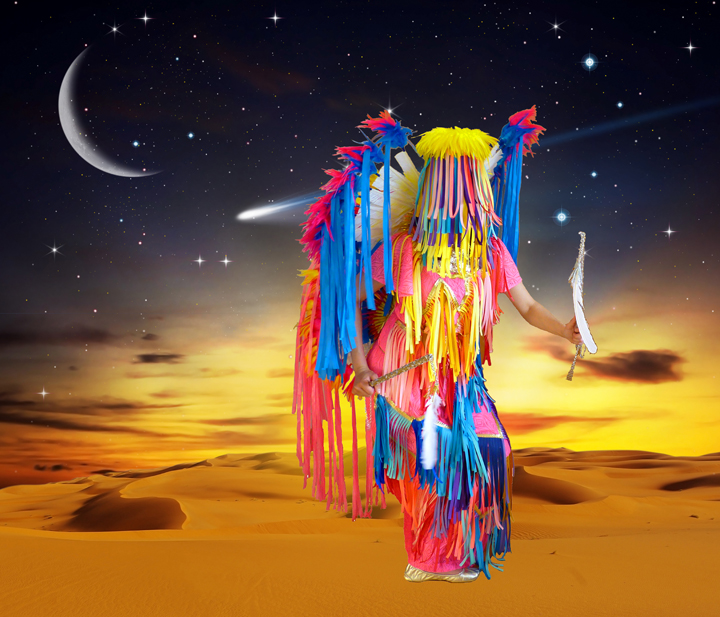 A vibrant, colorful figure stands in a desert landscape beneath a starlit night sky with a crescent moon and comet. The figure is draped in flowing ribbons and fringe in shades of hot pink, bright yellow, electric blue, and red, creating an explosive, dynamic silhouette against the golden sand and warm sunset horizon.