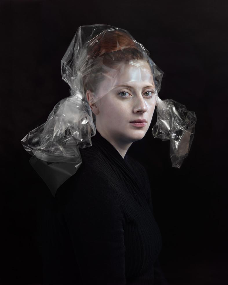 Portrait of a fair-skinned woman with reddish-brown hair styled close to her head, wearing a black ribbed garment against a black background. Transparent cellophane material is draped over her head and shoulders, creating translucent folds and reflective surfaces that frame her face and extend down her back.